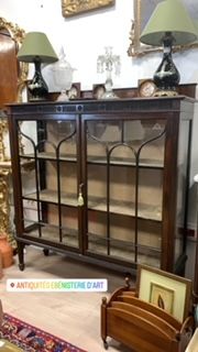 Vitrine ancienne en bois avec portes vitrées, deux lampes et objets décoratifs sur le dessus.