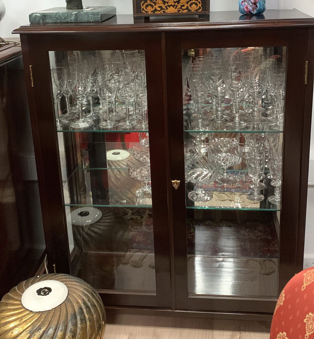 Vitrine en bois foncé à portes vitrées, remplie de verrerie. Quelques objets décoratifs sont visibles.
