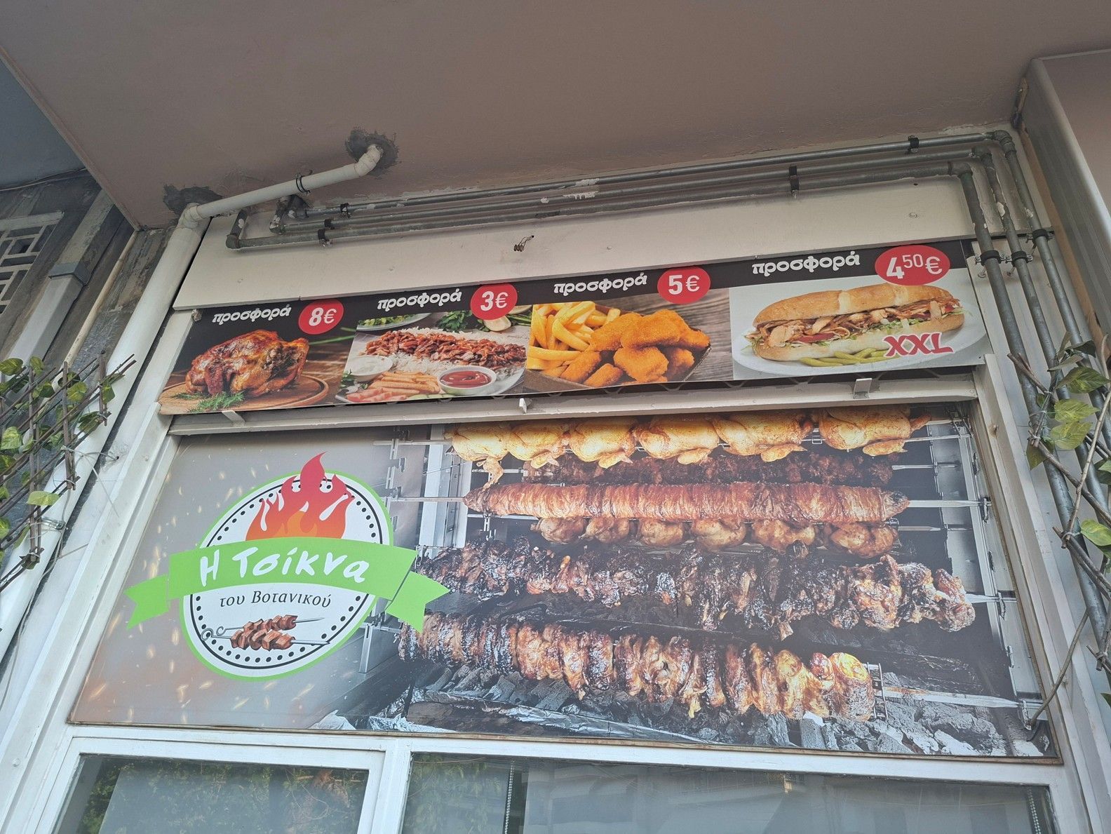 Βιτρίνα εστιατορίου με πανό τροφίμων και σουβλάκια, καθώς και μια πράσινη πινακίδα με το λογότυπο πάνω από την είσοδο
