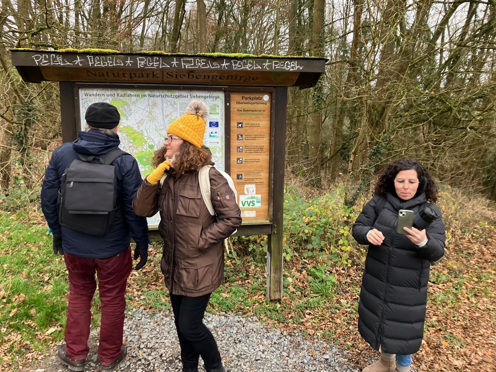 Drei Personen stehen an einem Wegweiser mit Karte und Hinweisschildern in einem Waldpark.