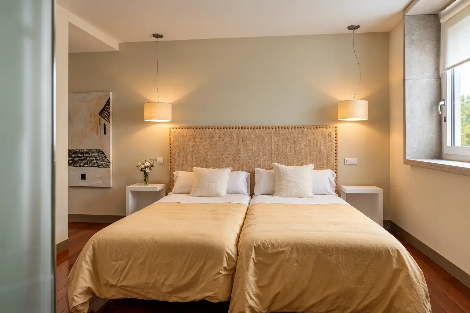 Habitación luminosa con cama, mesitas de noche, cuadro enmarcado, silla de madera y suelo de madera.
