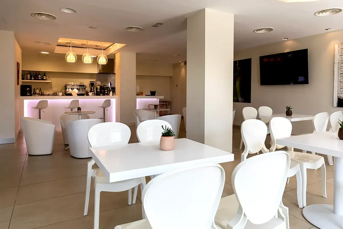 Interior moderno de cafetería con mesas y sillas blancas, una barra iluminada y un televisor montado en la pared.