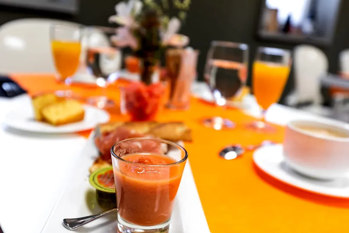 Mesa de desayuno con zumo de naranja, café, bollería y un vaso de batido sobre un mantel blanco