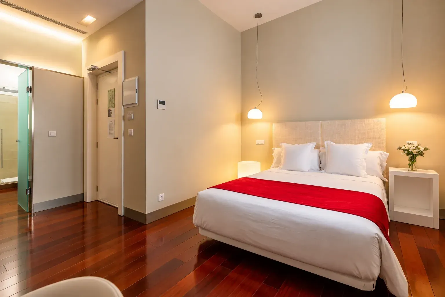 Habitación de hotel moderna con una cama blanca, una manta roja a los pies, lámparas colgantes y suelo de madera pulida