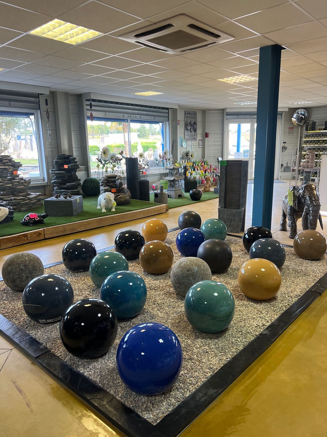 Présentation de sphères de pierre colorées sur un lit de gravier, à l'intérieur d'un magasin proposant des fontaines et autres éléments de décoration de jardin.