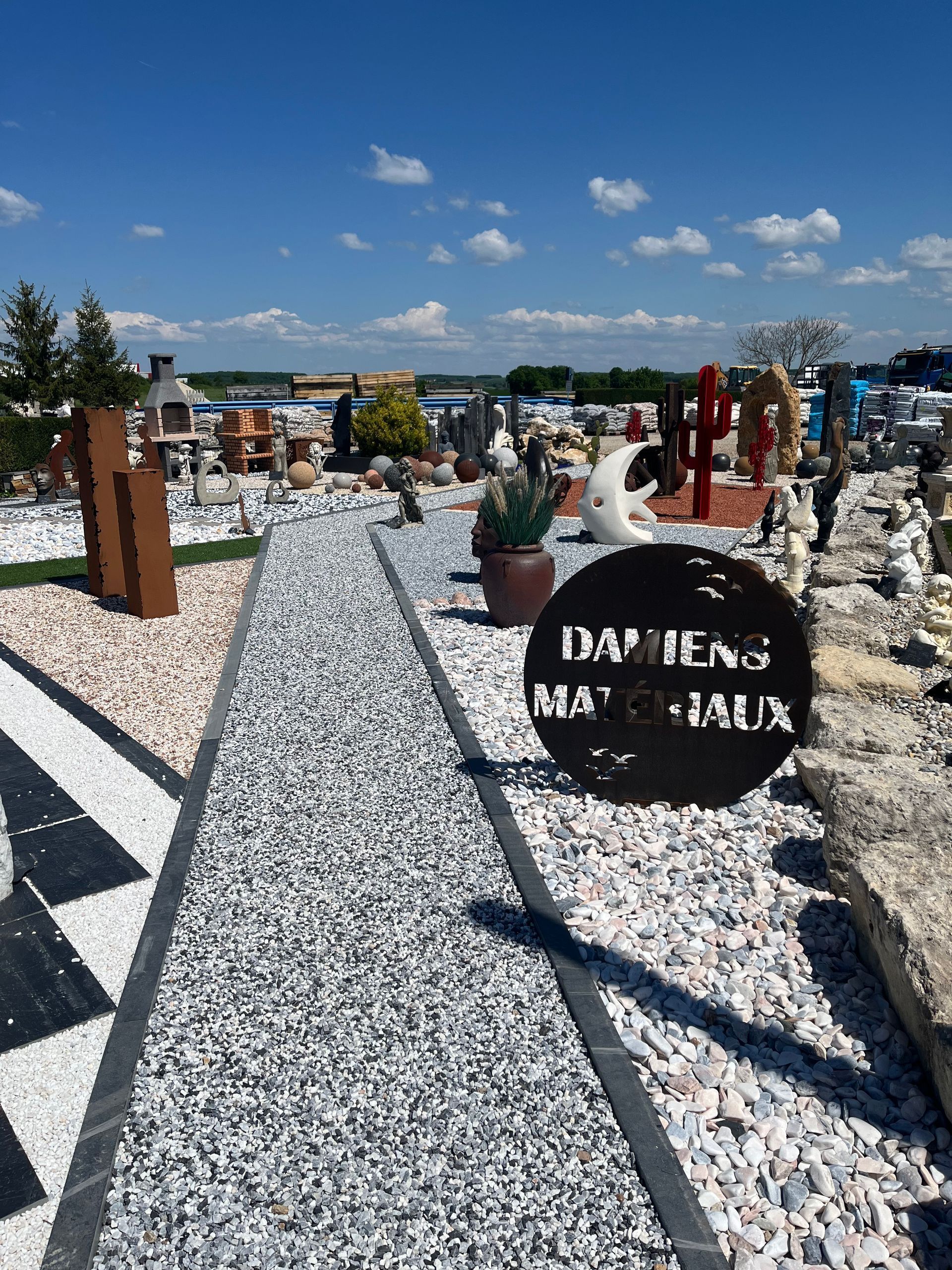Allée de pierres traversant une exposition extérieure de sculptures et d'éléments décoratifs de jardin sous un ciel bleu.