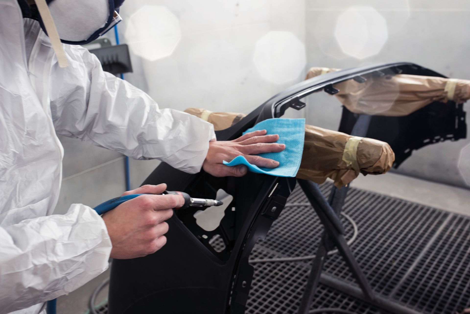 Une personne en combinaison de protection nettoie le pare-chocs d'une voiture dans une cabine de peinture.