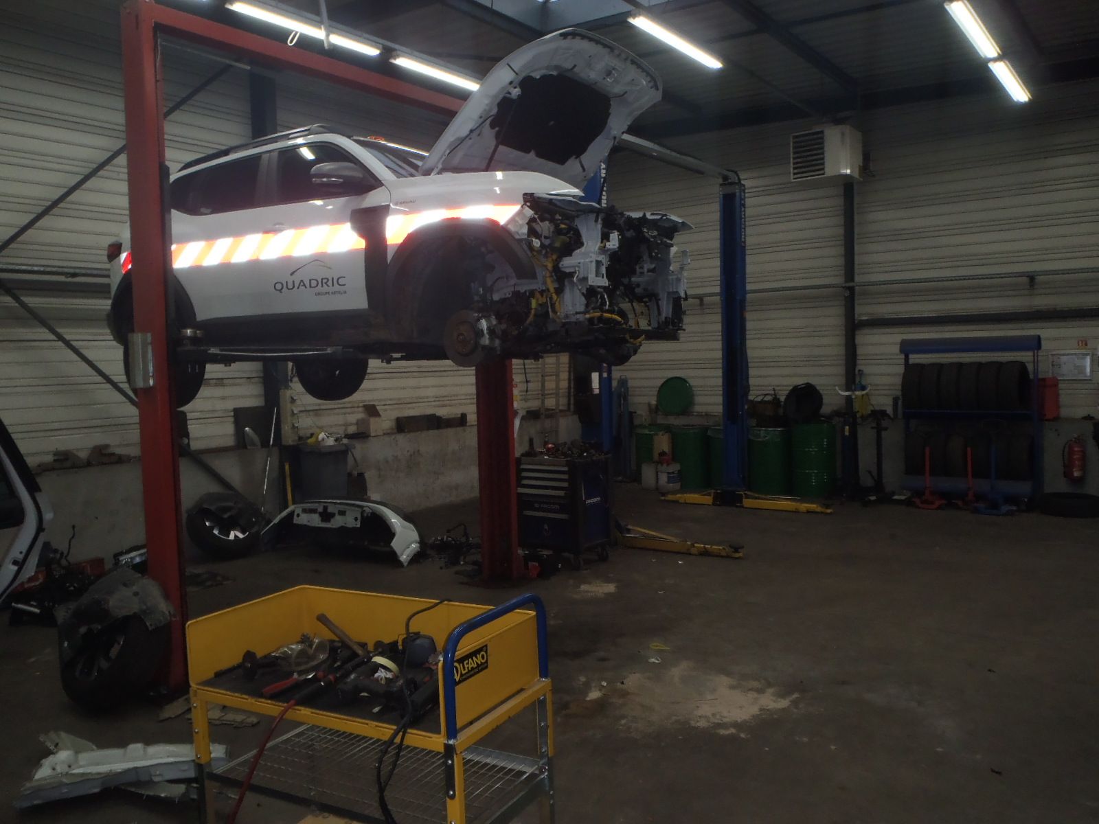 Voiture sur un pont élévateur dans un atelier de réparation ; capot ouvert, avant endommagé. Outils et pièces visibles.