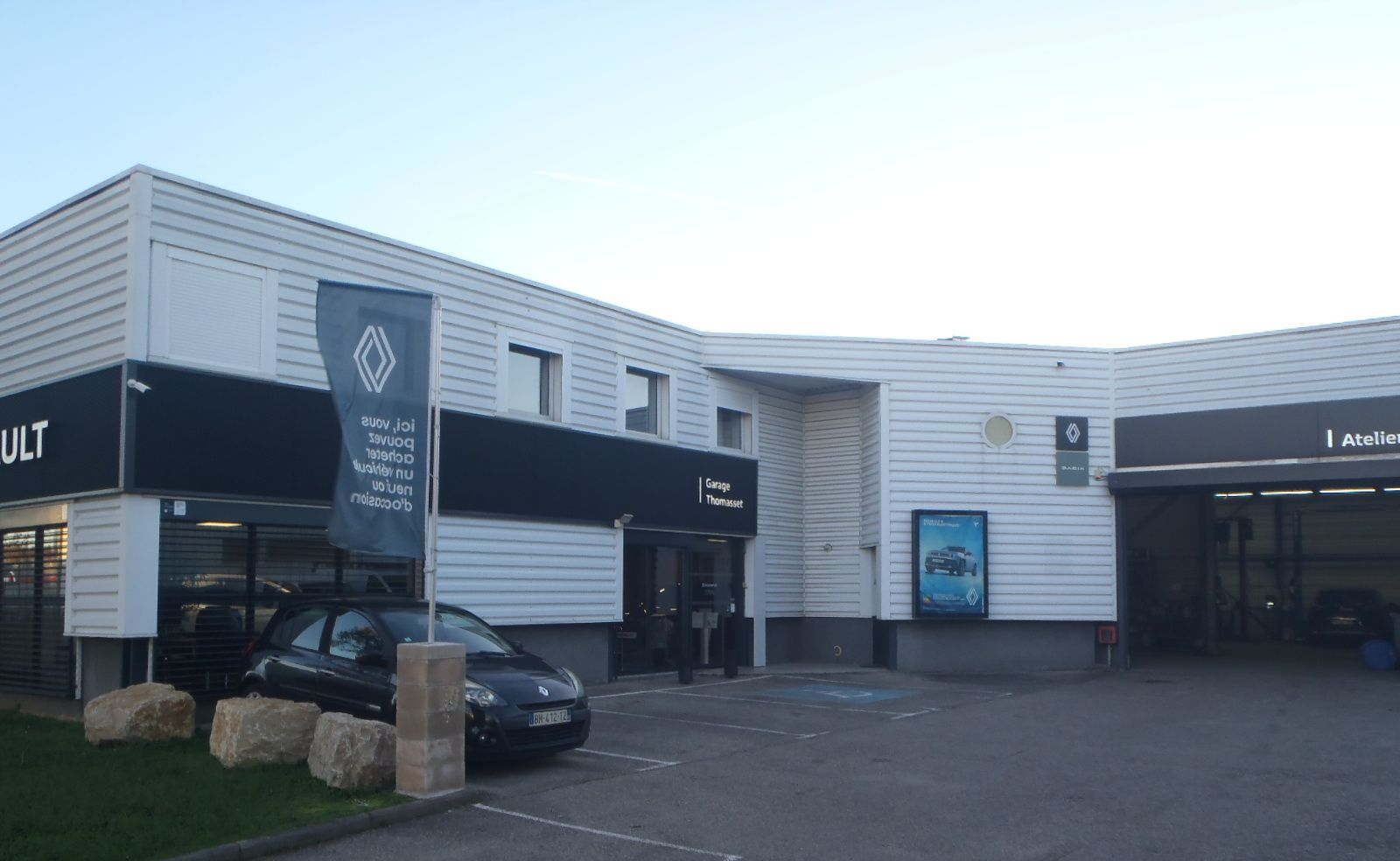 Concessionnaire Renault, vue extérieure. Voiture noire au premier plan. Bâtiment blanc avec enseignes bleues et noires.