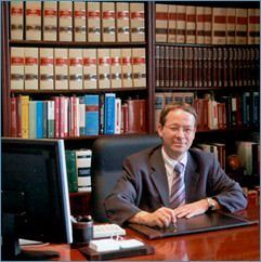 Un profesional sentado en un escritorio en un bufete de abogados, con estanterías repletas de volúmenes jurídicos al fondo.