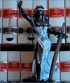 Una estatua de color bronce de la Dama de la Justicia sosteniendo una balanza y una espada, situada frente a una estantería llena de libros de leyes.