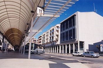Una terminal de autobuses al aire libre con un largo techo metálico arqueado, un autobús estacionado, un edificio moderno y coches bajo un cielo azul despejado.