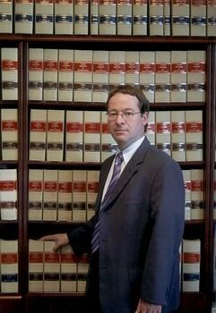 Una persona vestida con traje y gafas está de pie frente a una estantería llena de libros de derecho uniforme.