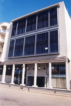 Vista desde un ángulo bajo de un edificio de varias plantas con una fachada que presenta grandes ventanas tintadas de oscuro sobre una columnata de pilares blancos.