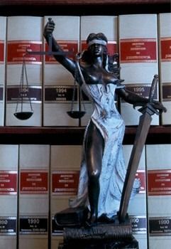 Una estatua de la Dama de la Justicia con balanza y espada, situada en el centro, frente a una estantería llena de libros de leyes.