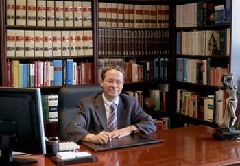 Una persona vestida de traje está sentada en un escritorio en un bufete de abogados, con estanterías llenas de libros de derecho y una estatua de la Dama de la Justicia al fondo.