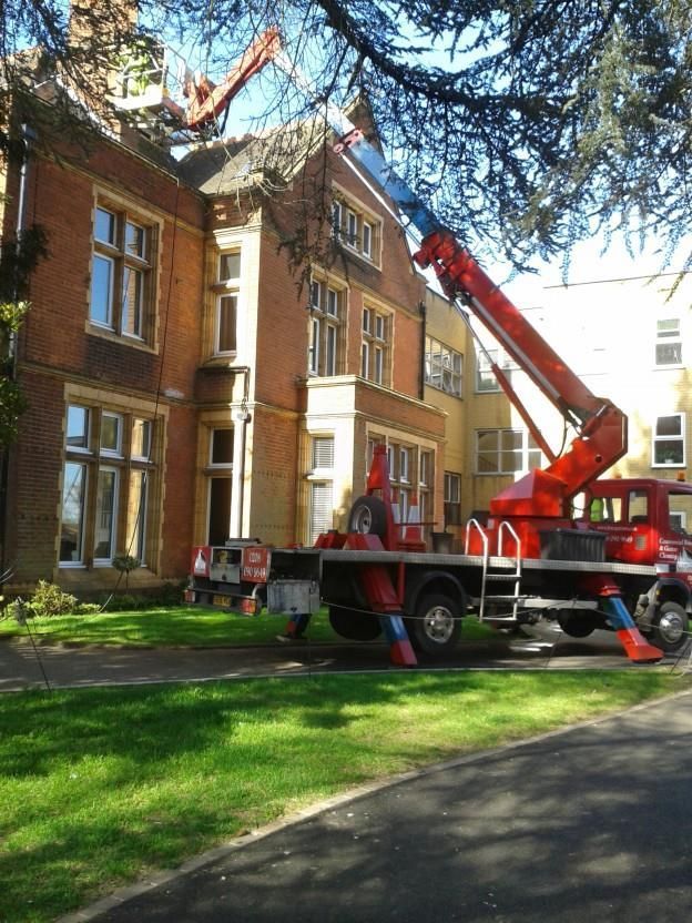 Red crane truck parked by a brick building, lifting equipment near the roof on a sunny day.