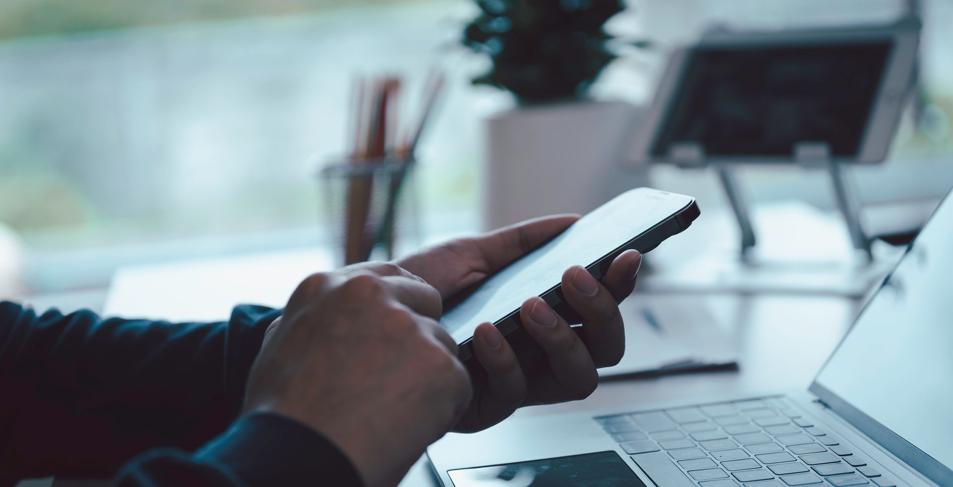 Des mains utilisant un smartphone, à proximité d'un ordinateur portable et d'une tablette, dans un environnement intérieur lumineux.