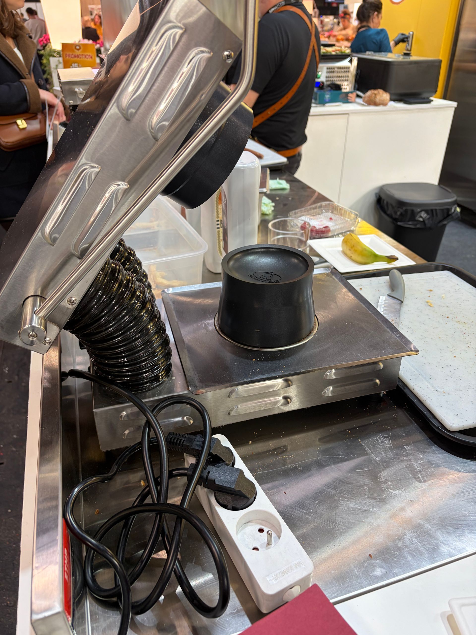 Poste de cuisson avec ustensiles en métal. Une casserole noire repose sur une surface argentée ; le câble électrique est visible. Des personnes sont visibles en arrière-plan.