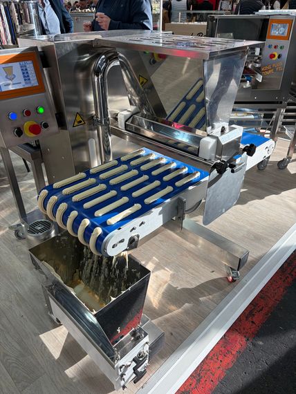 Vers la page boulangerie Machine de transformation alimentaire distribuant des bâtonnets de pâtisserie fourrés sur un tapis roulant. En acier inoxydable bleu, dans un environnement industriel.