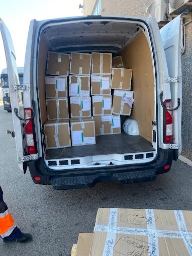 Vista trasera de una camioneta de reparto blanca con las puertas abiertas, cargada con cajas de cartón, al aire libre.