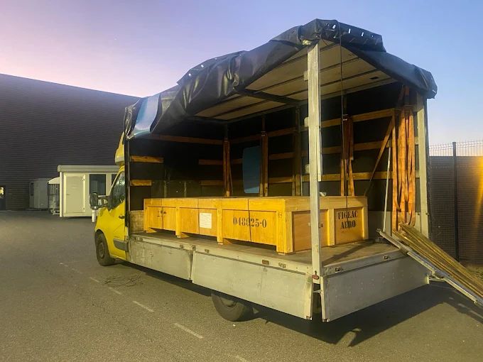Camión amarillo con cajas de madera cargadas en la caja, con una lona encima. Al aire libre.