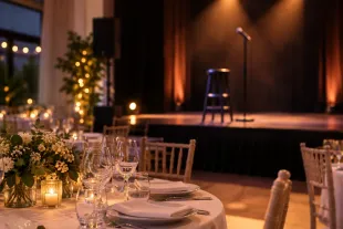 Elegante salón de banquetes con mesas iluminadas con velas y un escenario con micrófono y taburete.