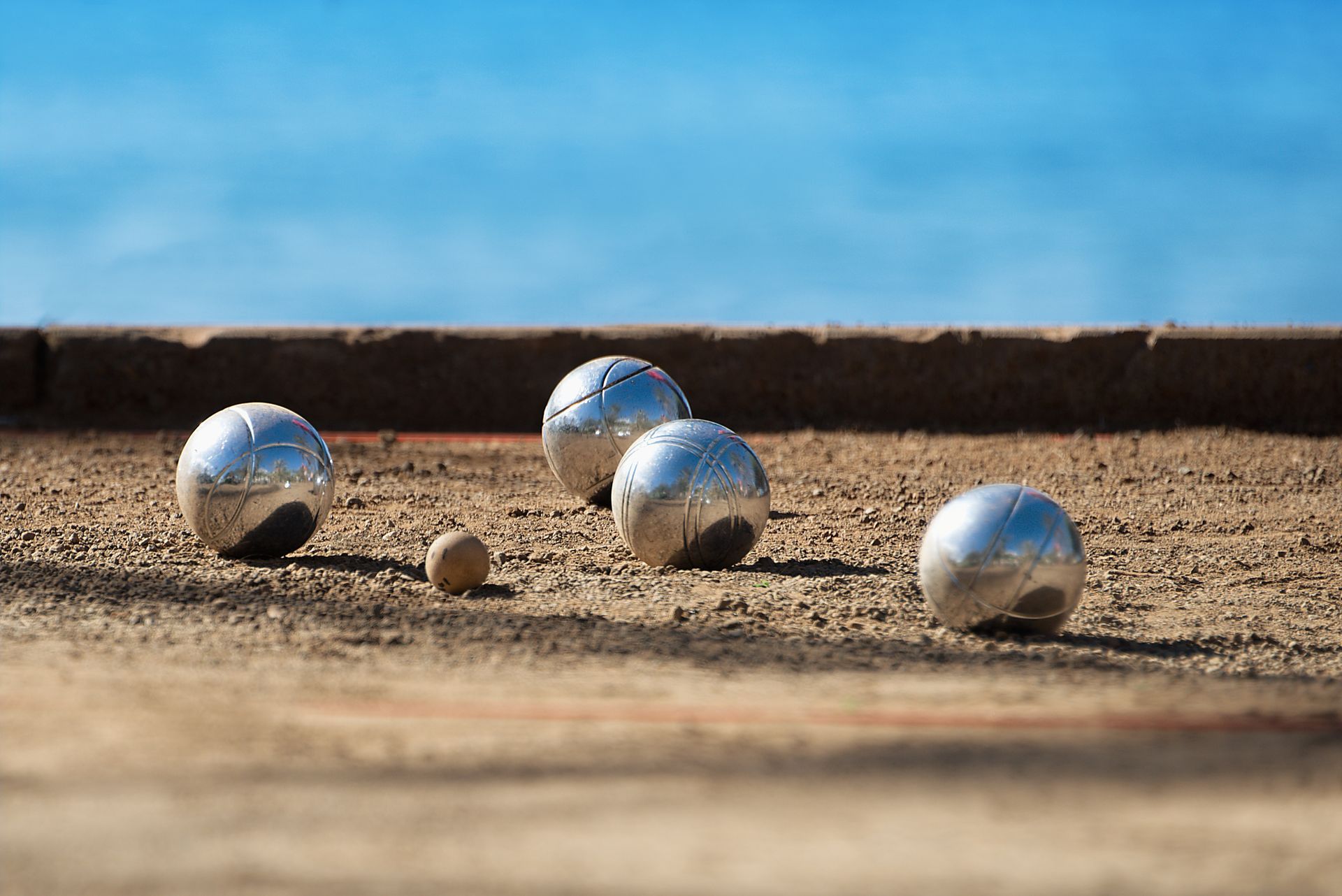 Boules de pétanque en métal et petit cochonnet sur un terrain de sable sous un ciel bleu.