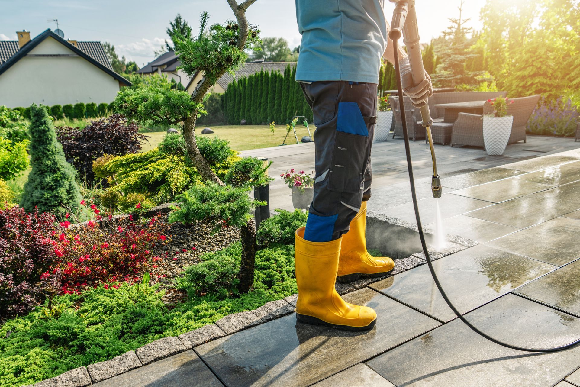 Une personne en bottes jaunes nettoie à haute pression une allée à côté d'un jardin paysager au coucher du soleil.