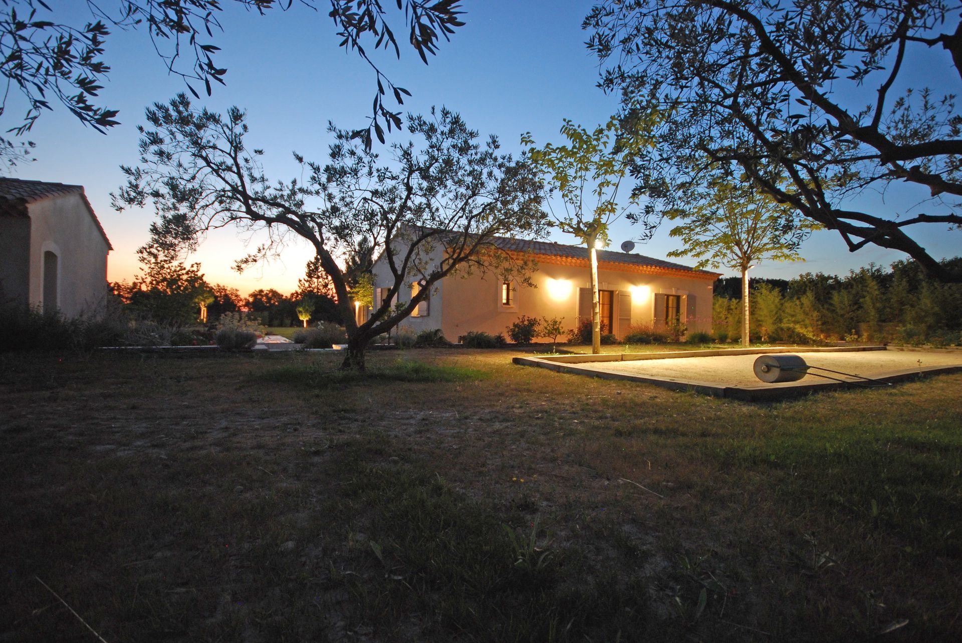 Maison faiblement éclairée au crépuscule, avec des lumières de porche scintillantes, des arbres et une cour au premier plan.
