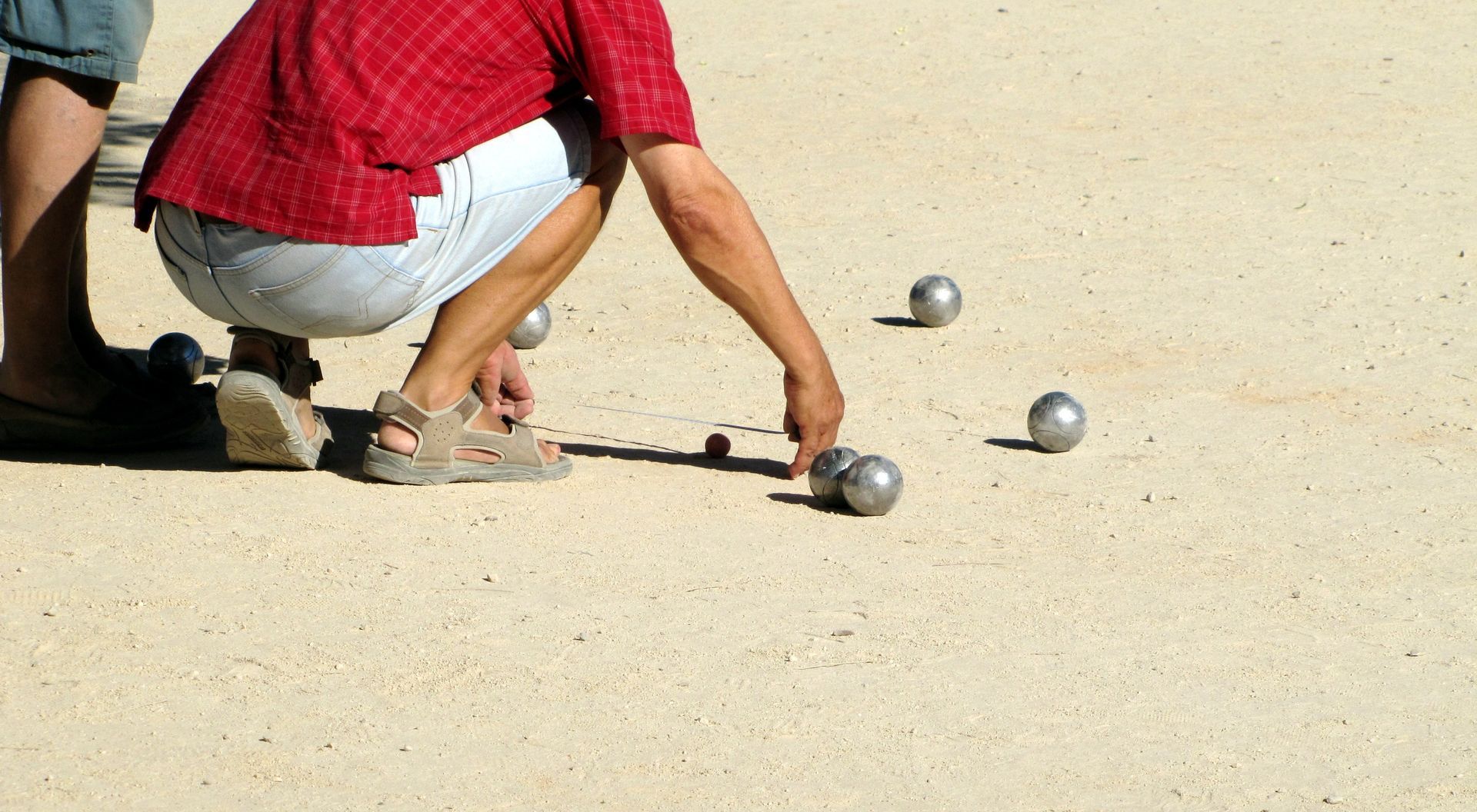 Une personne en chemise rouge est accroupie sur le sable, visant une boule de métal lors d'une partie de pétanque.