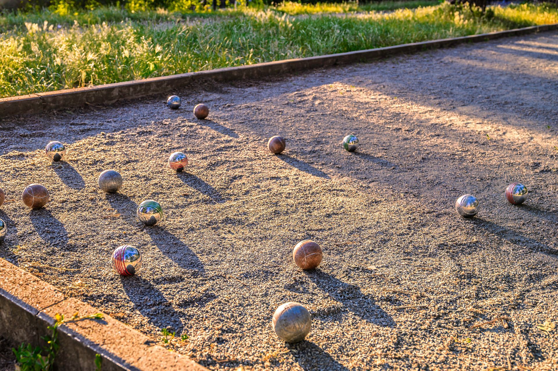 Des boules de métal éparpillées sur un terrain extérieur recouvert de gravier, baigné par un soleil chaud.