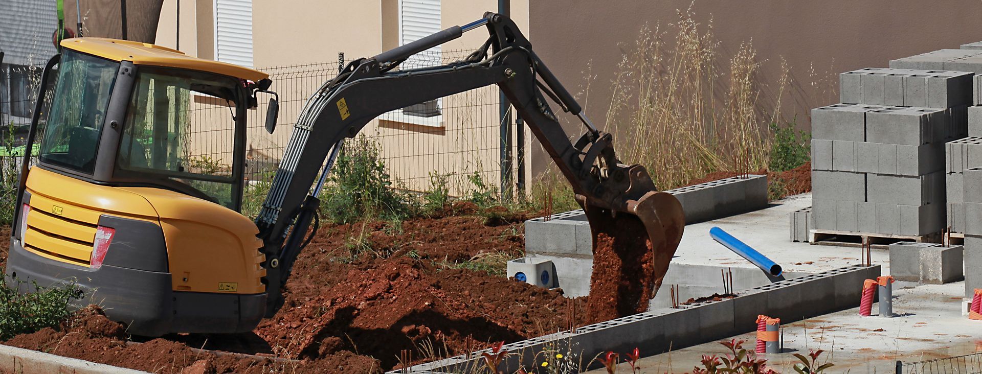 Une petite pelle mécanique creuse la terre à côté de blocs de béton empilés sur un chantier.