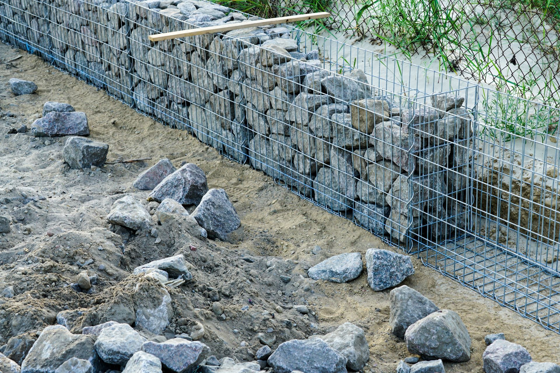 Mur de soutènement en pierre le long d'une pente boueuse, avec une clôture en grillage et des pierres éparses devant.