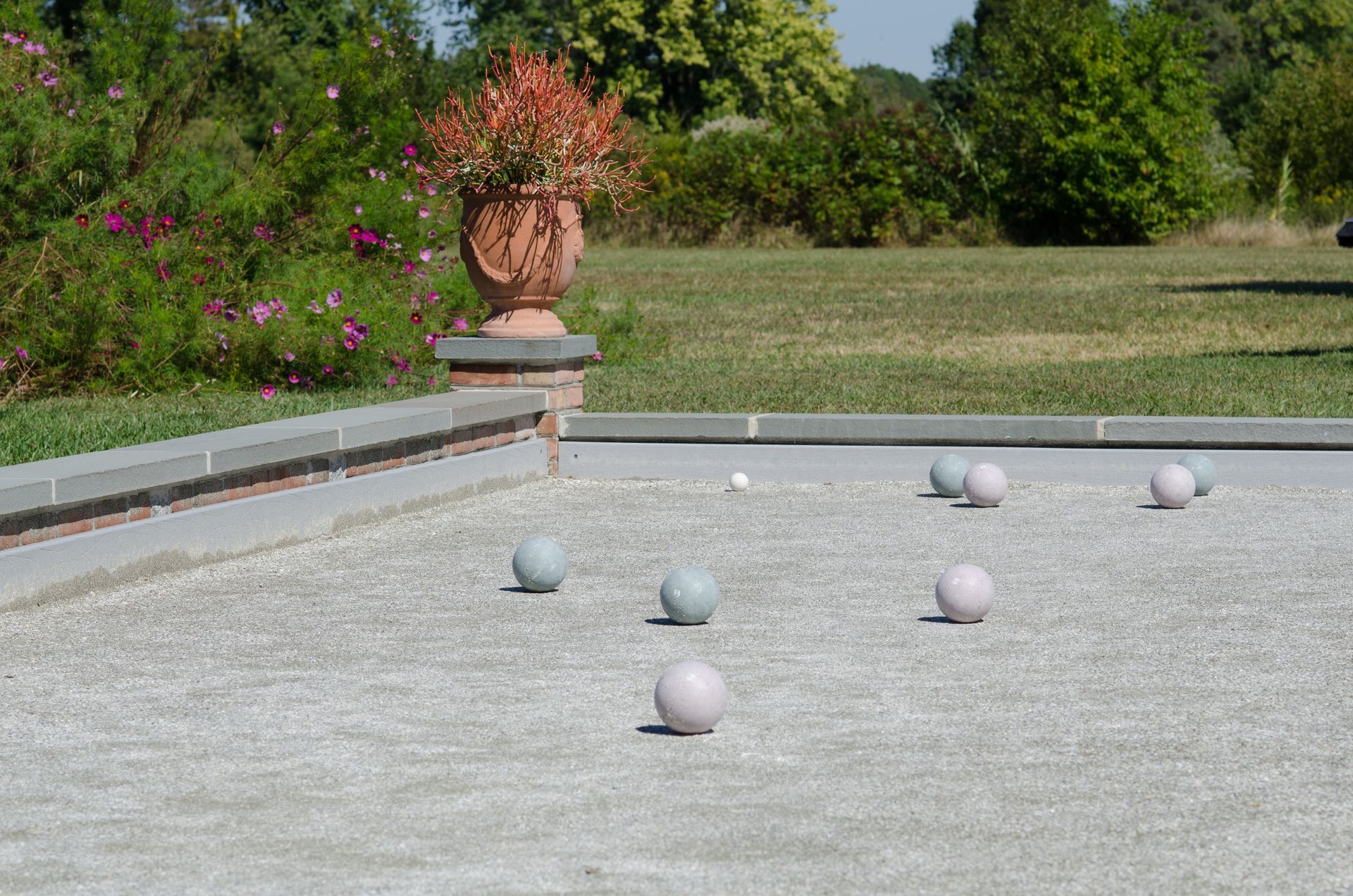 Plusieurs boules de pétanque pâles sur un terrain de gravier dans un jardin, avec une grande vasque et de la verdure en arrière-plan.