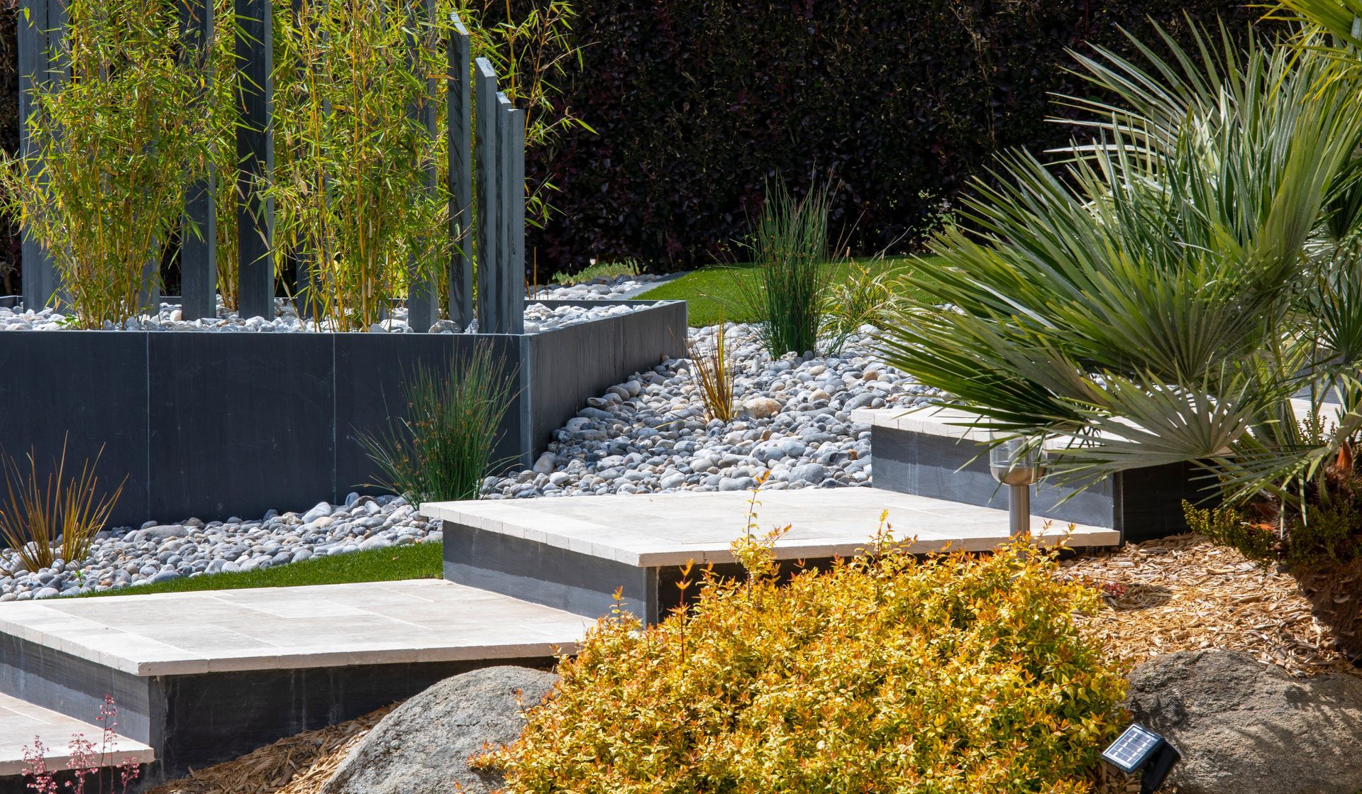 Jardin paysager moderne avec marches en béton, gravier blanc, palmiers et arbustes taillés.