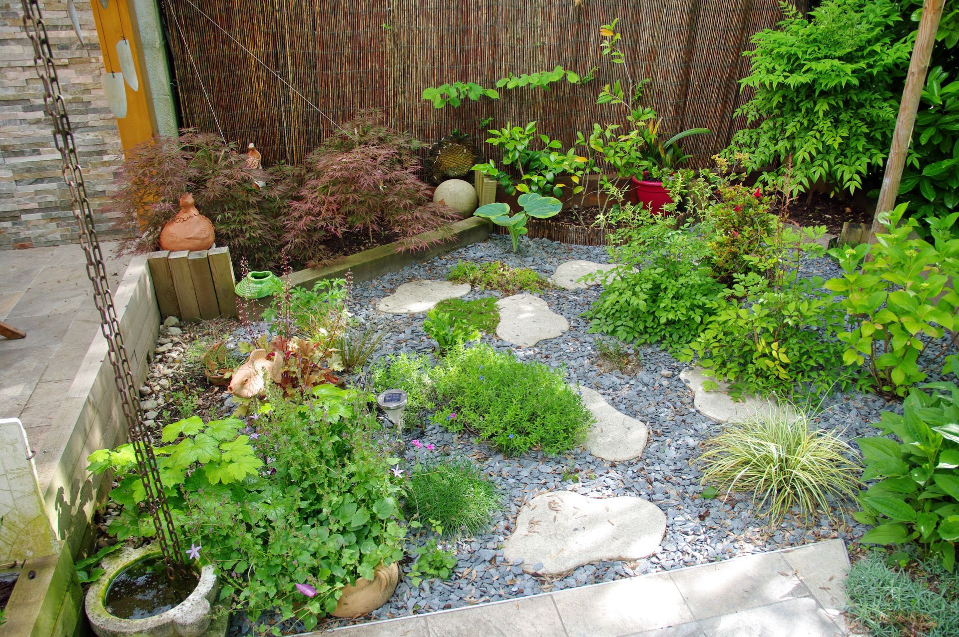 Petit chemin de jardin avec des dalles, du gravier et une végétation luxuriante longeant une clôture en bois.