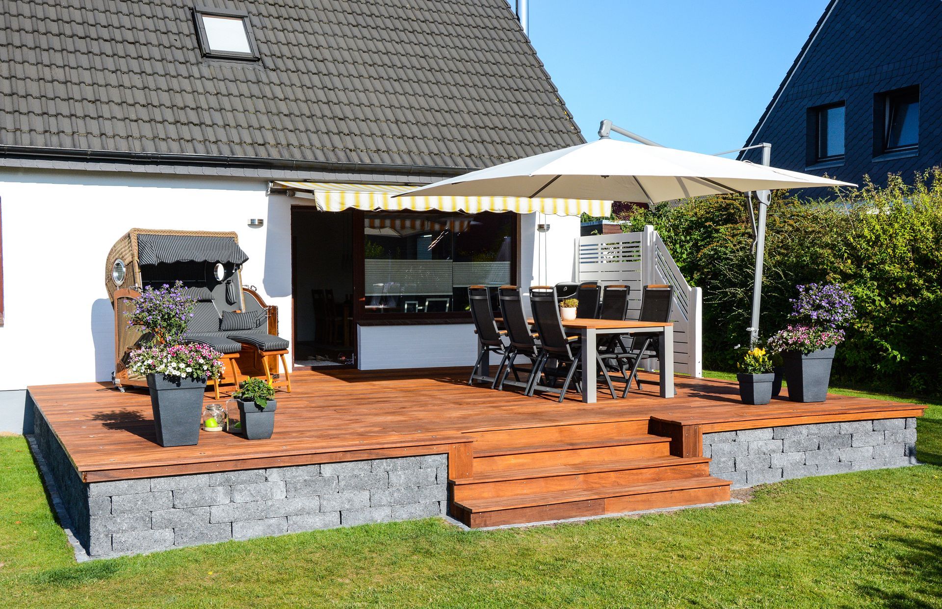 Terrasse arrière avec plancher en bois, parasol blanc, plantes en pot et sièges d'extérieur à côté d'une maison.