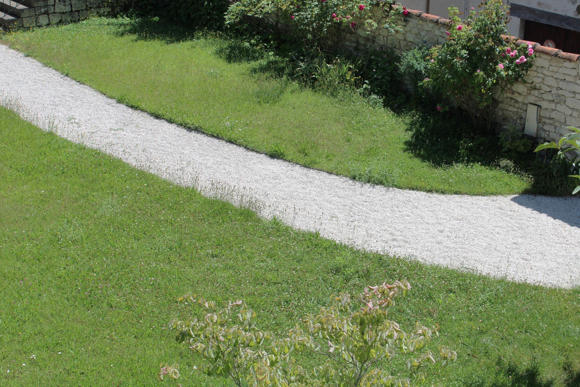 Un chemin de gravier blanc sinueux traverse une pelouse verte, longeant un muret de pierre agrémenté d'arbustes.