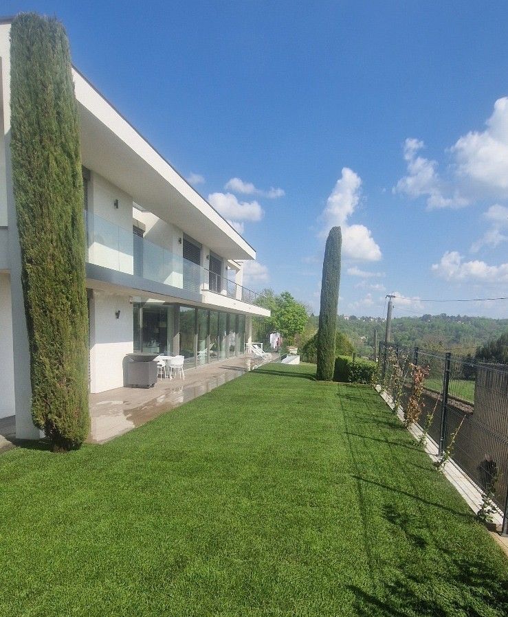 Maison blanche moderne avec pelouse verte, grands cyprès et vue sur une colline ensoleillée sous un ciel bleu.