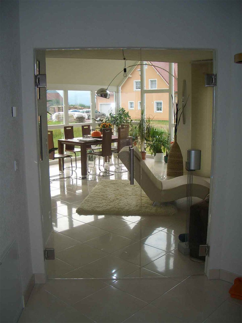 Durch eine Glastür gelangt man in den Essbereich mit Tisch, Stühlen, Pflanzen und Sofa. Heller, sonniger Innenraum.