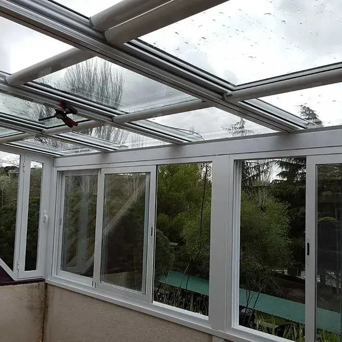Terraza soleada con techo de cristal, marcos blancos y ventanas con vista a una exuberante vegetación.