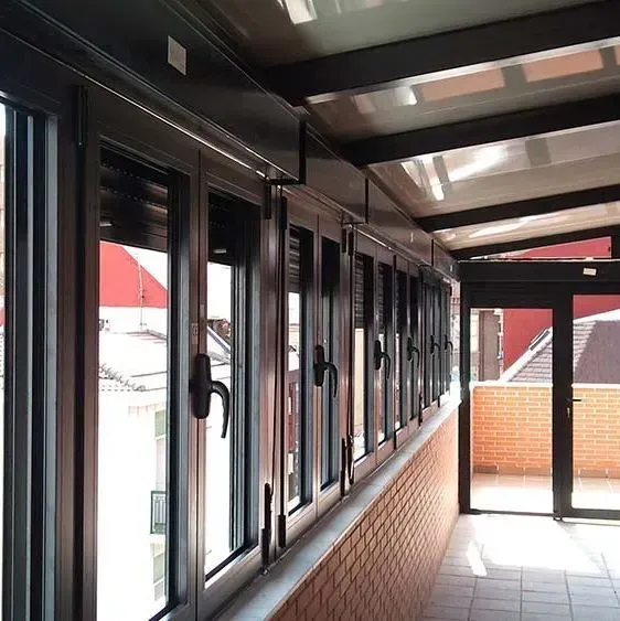 Hilera de ventanas con marcos negros sobre una pared de ladrillos; un techo de cristal cubre el balcón cerrado.