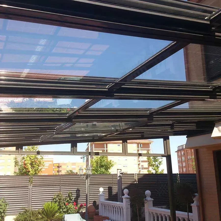 Pérgola de vidrio y metal sobre un patio; día soleado, cielo azul visible a través de los paneles de vidrio.