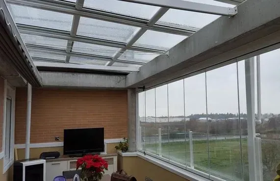 Terraza acristalada con paredes y techo de cristal, con vistas a un paisaje verde. En el interior hay un televisor y muebles.