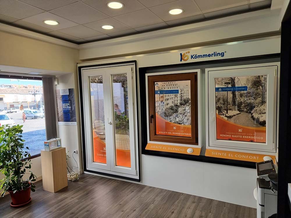 Interior de una sala de exposición de escaparates; se exhiben escaparates y carteles promocionales, una planta y una ventana abierta con vista a la calle.