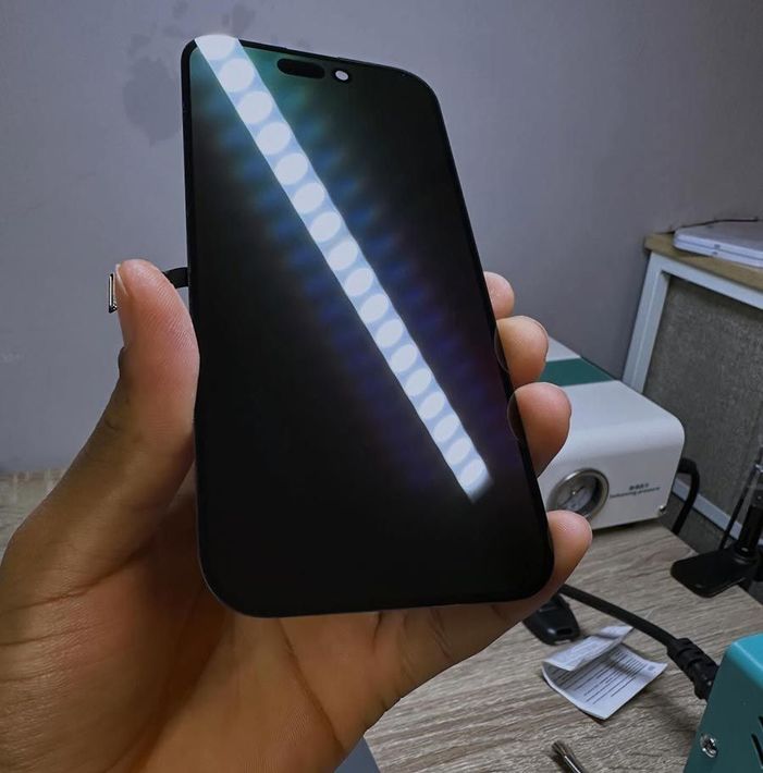 Una mano sostiene un smartphone negro con un reflejo diagonal brillante en una habitación