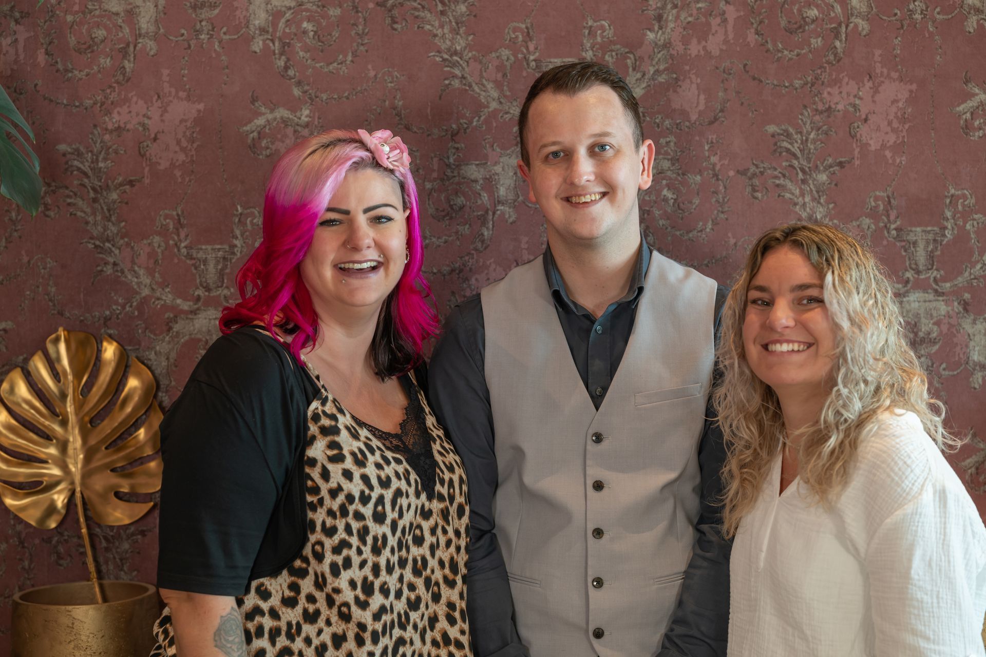 Drei Menschen lächeln in die Kamera. Der Mann links in Schwarz, der Mann in der Mitte in Weiß, die Frau rechts mit rosa und lila Haaren.