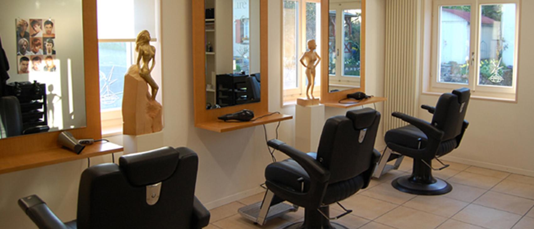 Innenraum eines Friseursalons mit drei Friseurstühlen, großen Spiegeln und Holzskulpturen.