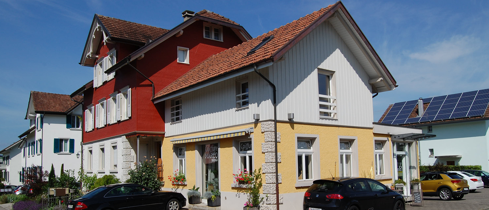 Gebäude mit roten, weißen und gelben Wänden unter blauem Himmel. Davor parken Autos.